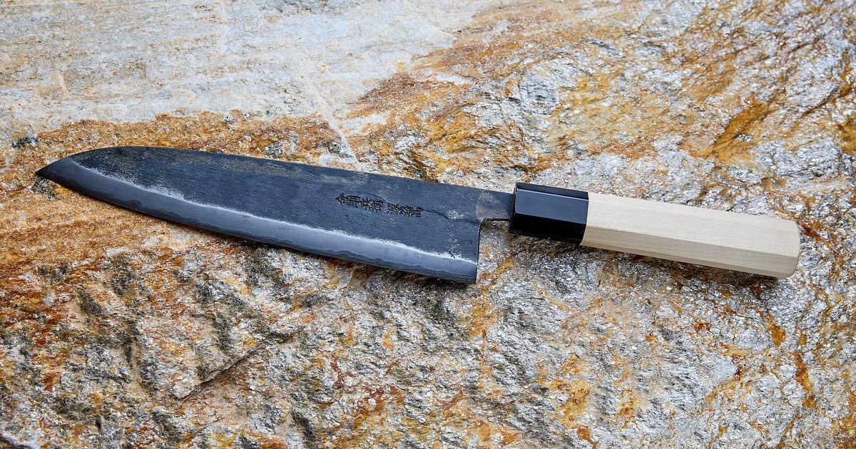 A hand-forged Japanese chef's knife with a dark, rough-finished blade, a polished edge and a pale wooden handle, lying on a rock