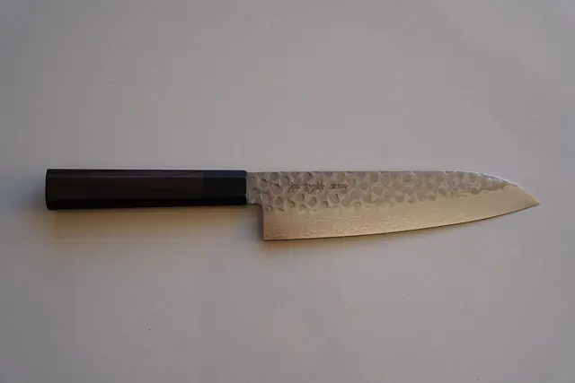 A santoku knife with a hammered pattern on the blade and a dark wooden handle, on a grey background