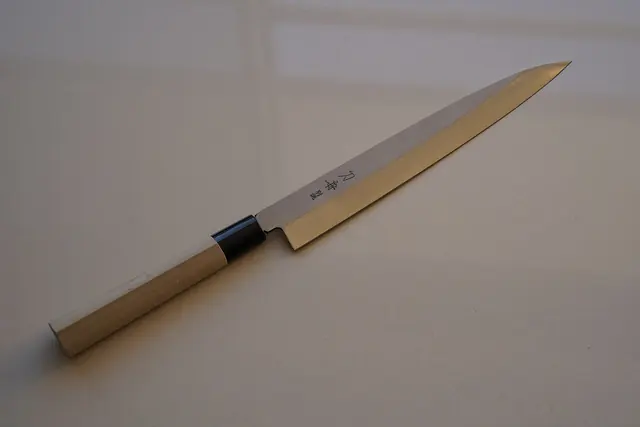 A long, narrow sashimi knife with a pale wooden handle, on a grey background