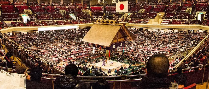 A packed arena seen from high red seats, with the ring and its hanging roof far below and a Japanese flag on the far wall