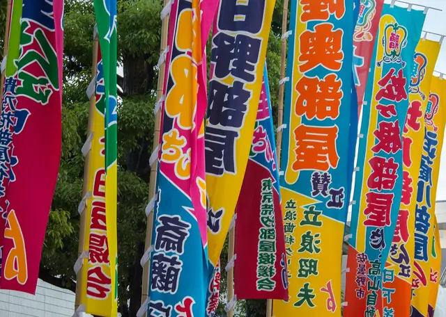 Tall cloth banners in pink, blue, yellow and green, each painted with a name in large black characters, lined up under trees