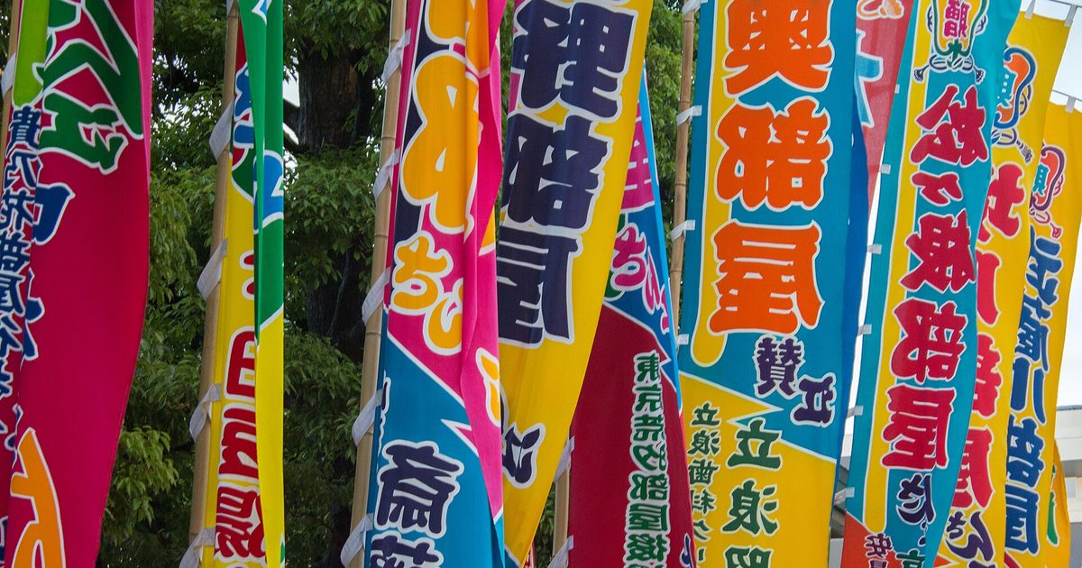 Tall cloth banners in pink, blue, yellow and green, each painted with a name in large black characters, lined up under trees