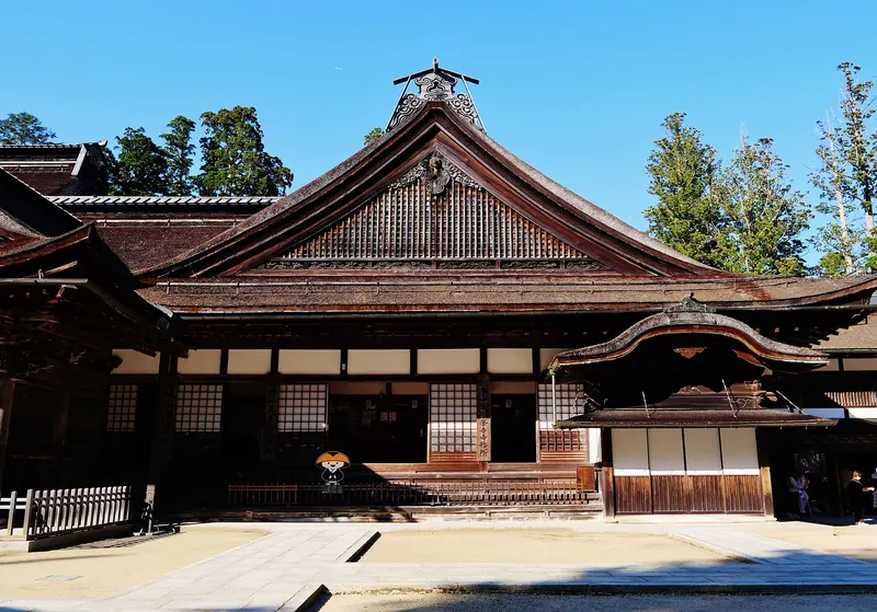A large wooden temple hall with a sweeping brown roof and a curved entrance porch under a blue sky