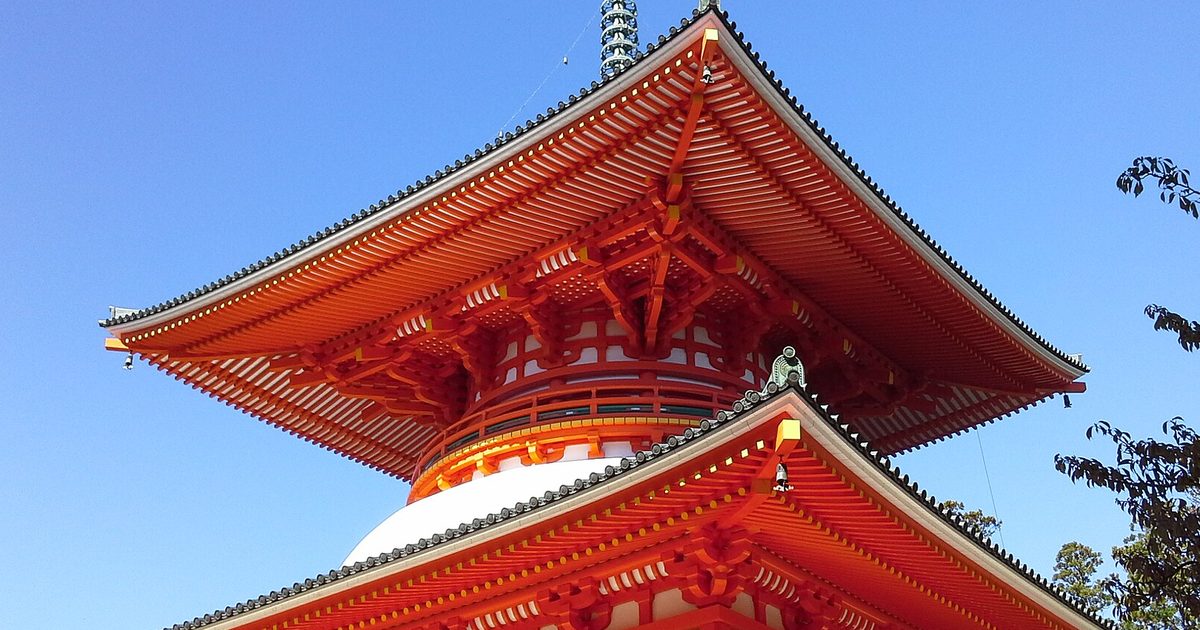 The vermilion upper roof and white dome of a two-storey pagoda against a clear blue sky, with a pine branch on the right