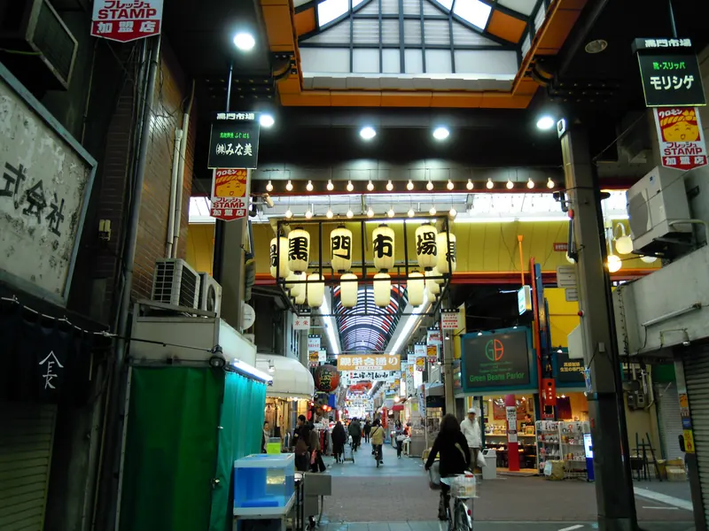 The arcade entrance of Kuromon Market with a row of lanterns, a cyclist seen from behind and shoppers