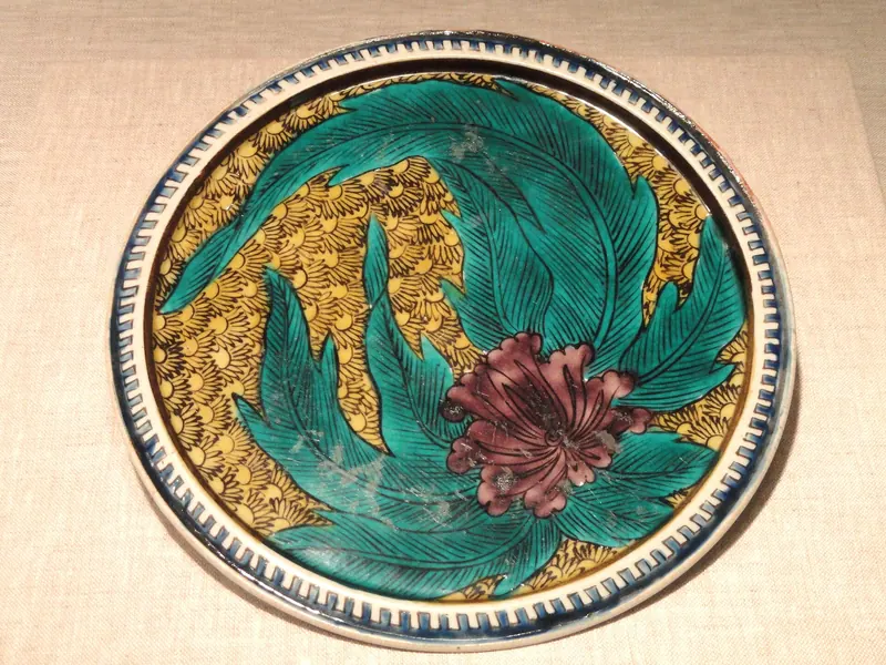 A round plate painted with large green leaves and a red flower over a yellow patterned ground