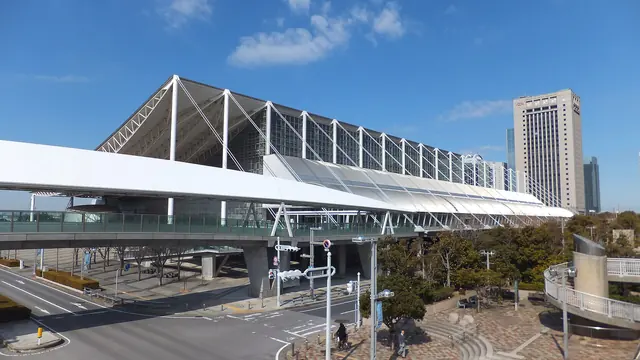 A large exhibition hall with a roof hung from white masts and cables, and a covered footbridge in front, under a blue sky