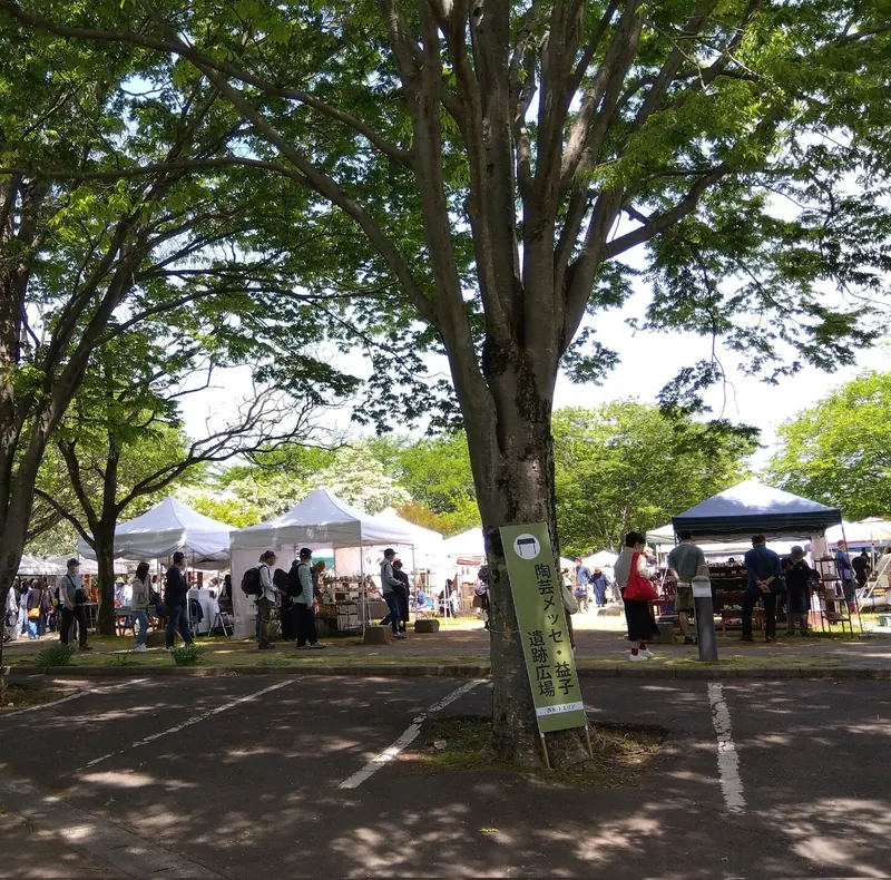 White market tents under tall trees, with shoppers small in the distance and a car park in front