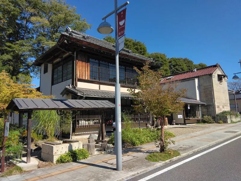 A two-storey wooden building with a tiled roof and a white storehouse beside it, set back from a quiet road with a lamp post