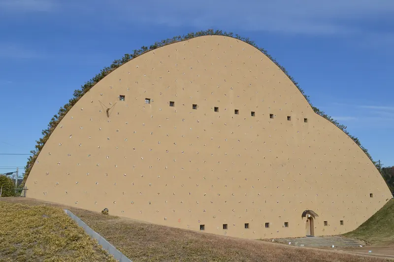 A large earth-coloured building shaped like a rounded hill, dotted with small square windows and with grass on top, under a blue sky