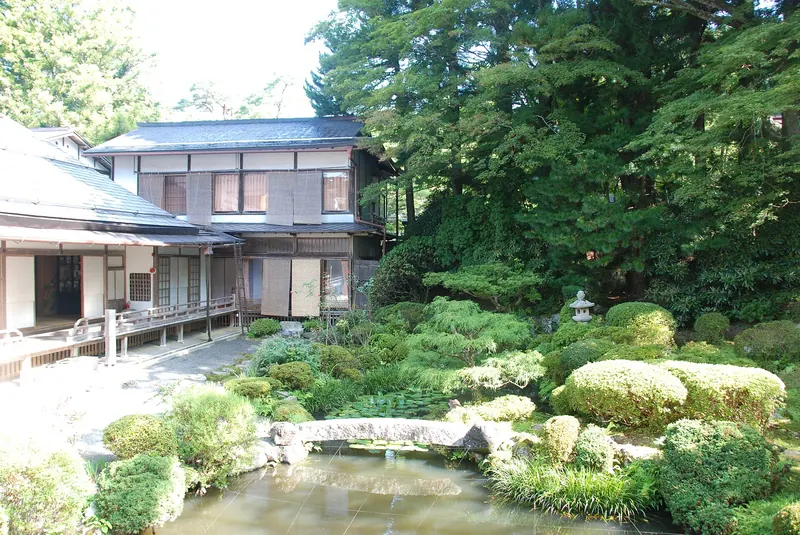 A temple garden of clipped shrubs, a pond and a stone lantern in front of a two-storey wooden building among tall trees
