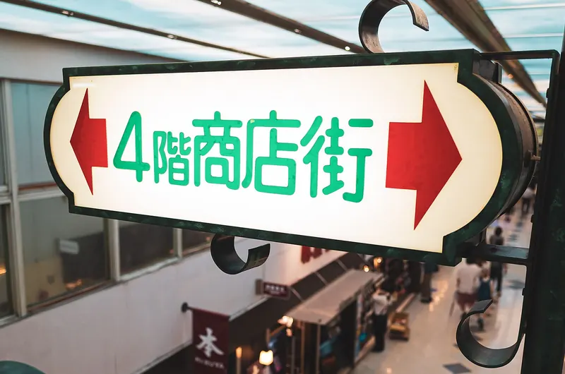 An illuminated hanging sign with green Japanese lettering and two red arrows, above an indoor shopping corridor seen from the floor above