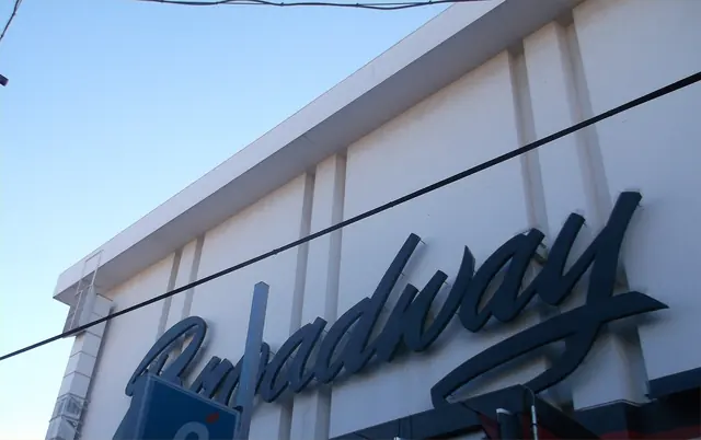 Large dark blue script letters spelling Broadway across the white upper wall of a building, against a blue sky with overhead wires in front
