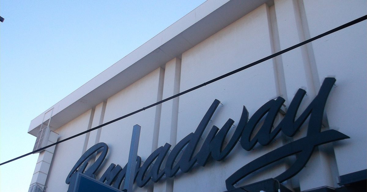 Large dark blue script letters spelling Broadway across the white upper wall of a building, against a blue sky with overhead wires in front