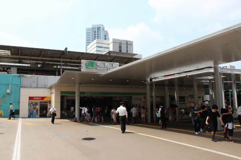 A long white canopy over the north entrance of a station with a green JR sign, and people crossing the open plaza in front