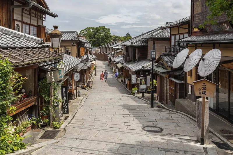 A stone-paved lane lined with wooden houses, with three white paper umbrellas on a shopfront on the right