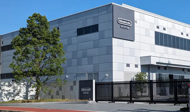 A grey box-shaped building with the Nintendo Museum logo on its wall, behind a black gate and a large tree under a blue sky