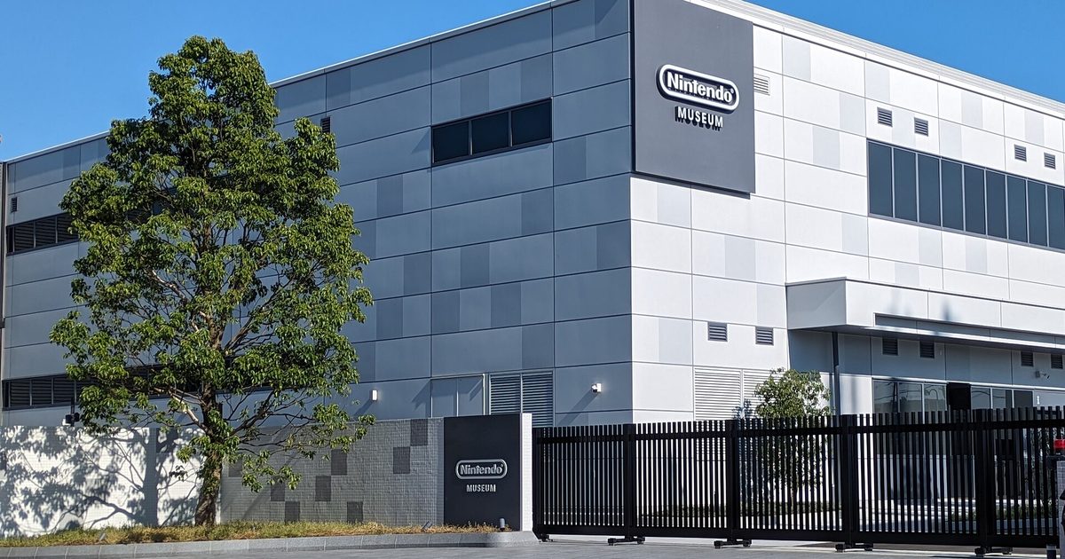 A grey box-shaped building with the Nintendo Museum logo on its wall, behind a black gate and a large tree under a blue sky