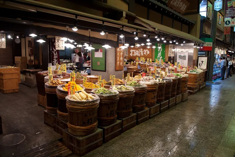 A pickle shop with wooden tubs heaped with vegetables under the arcade lamps