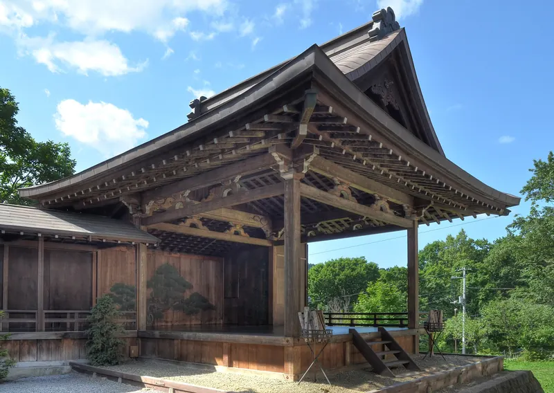 An outdoor wooden noh stage with a large tiled roof and a pine tree painted on the back wall