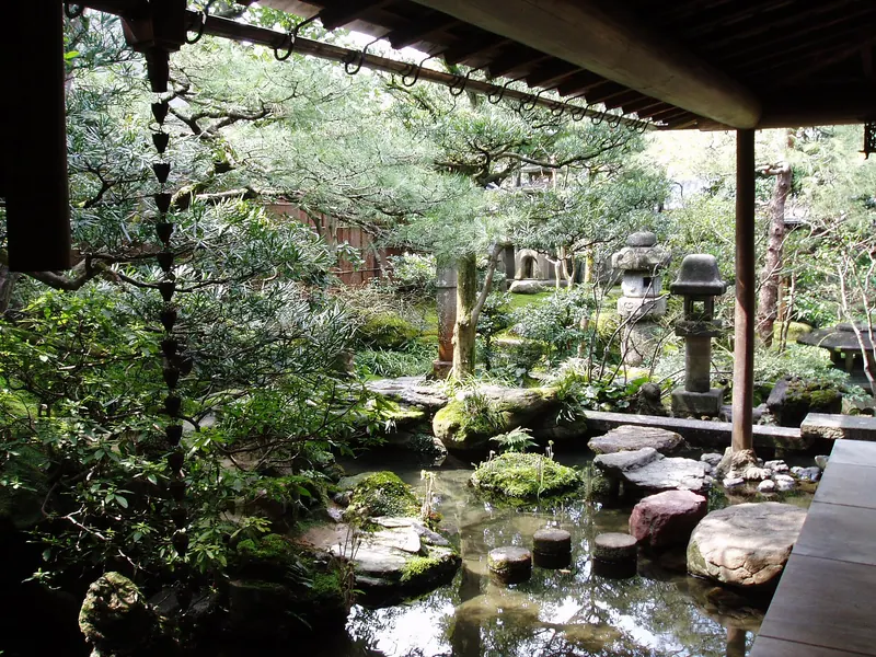 A small garden of pond, stepping stones, stone lanterns and pines seen from under the eaves of a wooden house