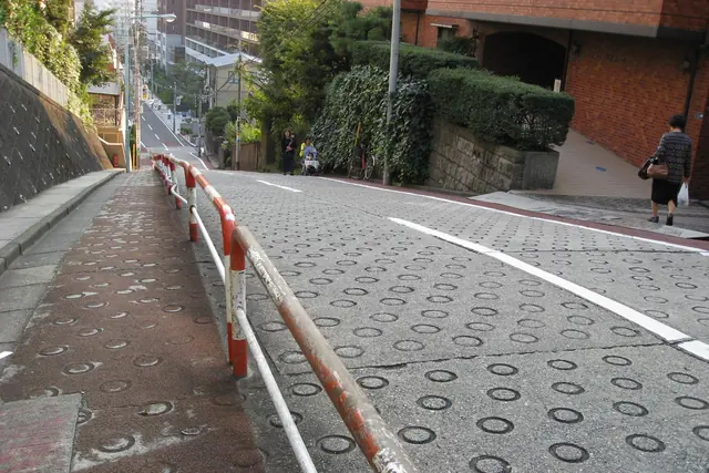 A steep road dropping away from the camera, its surface covered in round anti-slip rings, with red and white handrails along the side and a brick building on the right