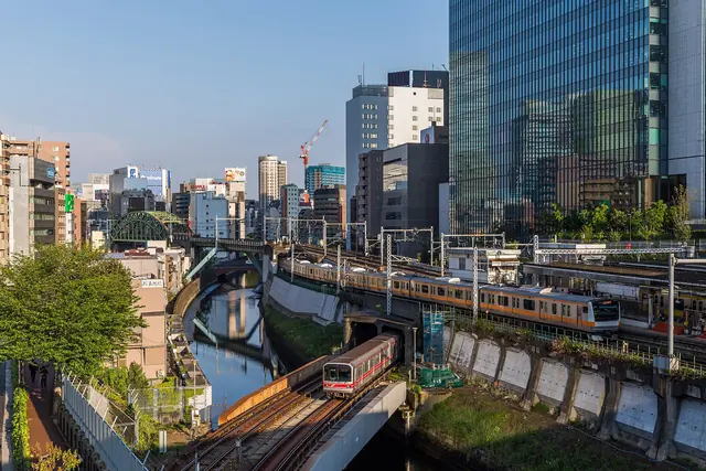 A river in a deep cutting seen from above, with a metro train crossing it on a low bridge, an orange-striped train on the bank and glass office towers behind