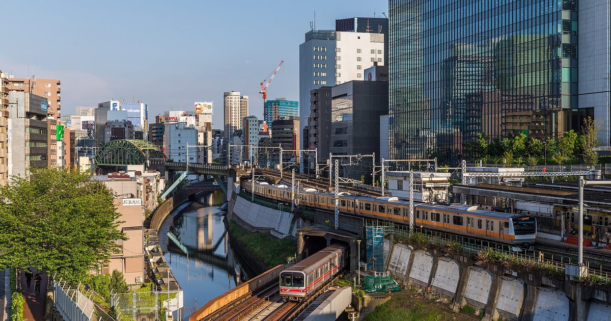 A river in a deep cutting seen from above, with a metro train crossing it on a low bridge, an orange-striped train on the bank and glass office towers behind