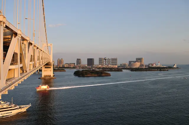 The side of a bridge on the left, small wooded islands and waterfront buildings across the water, and a boat leaving a wake in low sun