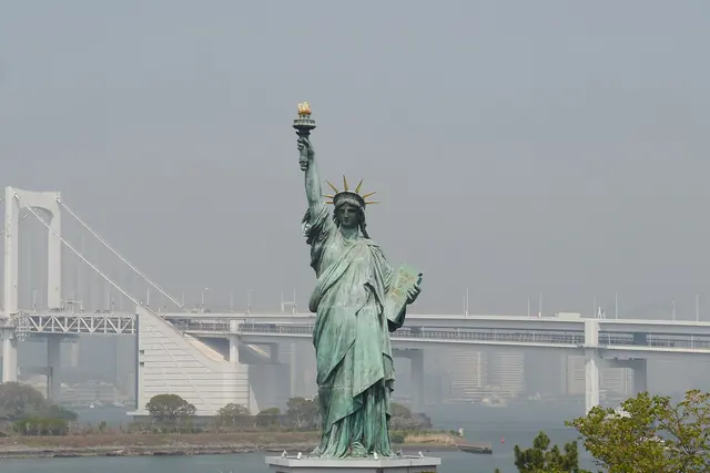 A pale green statue holding up a torch on a stone base, with a suspension bridge behind it in haze