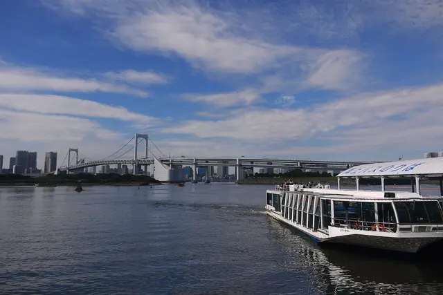A glass-sided boat at a roofed pier on a wide bay, with a suspension bridge in the distance under a blue sky with thin cloud