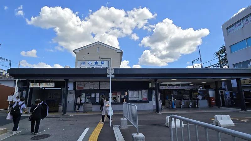 The low station building of Kintetsu Ogura Station with ticket gates and a station sign under a blue sky with white clouds