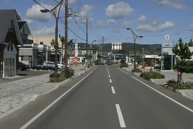 A wide straight street with paved footpaths, low shops and houses, street lamps and a few parked cars under scattered clouds