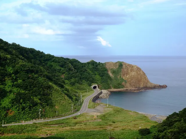 A road curving along a bay towards a tunnel under a rocky headland, with green slopes on the left and the sea beyond