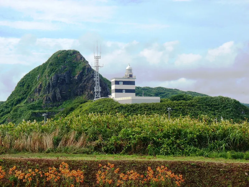 A white lighthouse beside a radio mast below a steep green rocky hill, with tall grass and orange flowers in the foreground