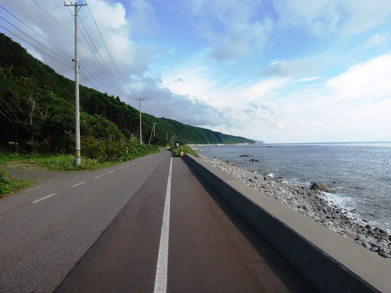 A two-lane road with a red-surfaced strip beside a low sea wall and a pebble beach, with a green slope on the left
