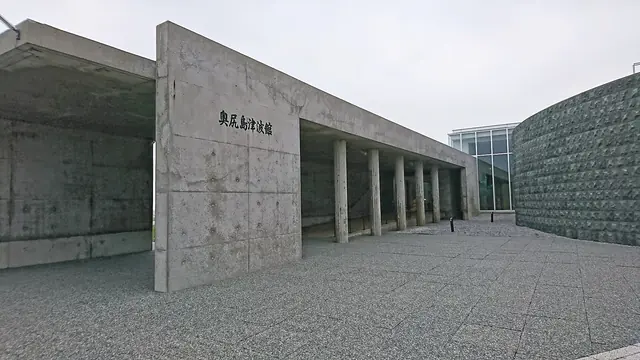 A bare concrete building with the museum's name in Japanese on a tall wall, a row of square columns and a curved stone wall to the right