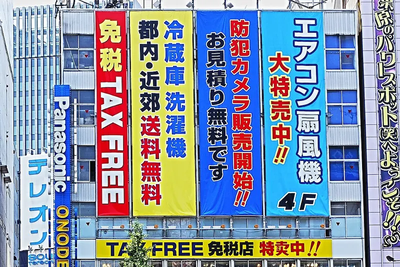 A shopfront covered in tall red, yellow and blue banners, one reading TAX FREE, above a yellow strip reading TAX FREE in English and Japanese, with a vertical ONODEN sign at the left