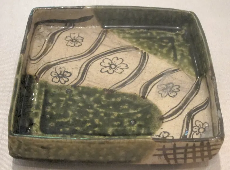 A square tray with patches of deep green glaze and brown painted flowers and wavy lines on a cream ground