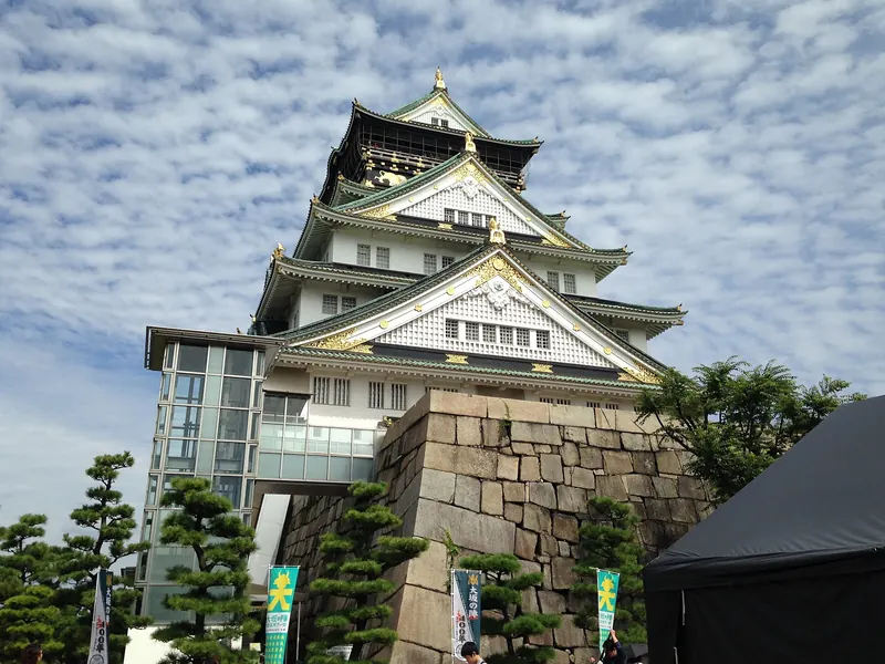The main tower of Osaka Castle with green roofs and gold ornaments seen from below above a stone wall and pine trees
