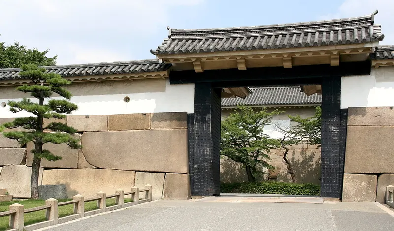 A black wooden gate with a tiled roof between huge stone blocks and a white wall, with a pine tree