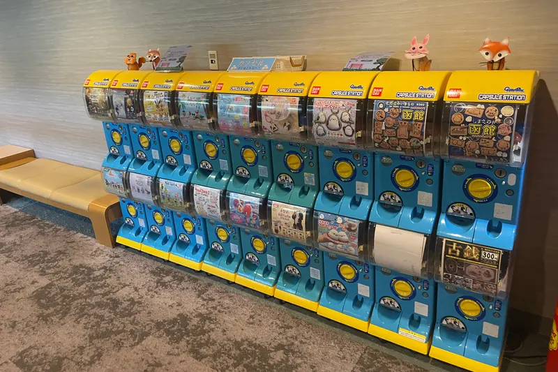 Two rows of blue-and-yellow capsule-toy machines along a wall, each showing a picture card of the toys inside, with small animal figures standing on top