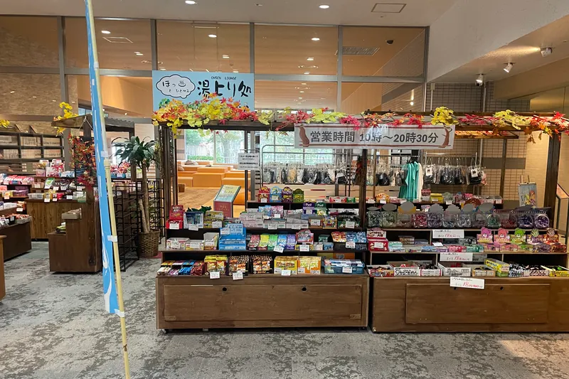 Low wooden stalls stacked with boxes of small sweets in front of a lounge entrance with a dark sign reading ONSEN LOUNGE, and a banner giving opening hours of 10:00 to 20:30