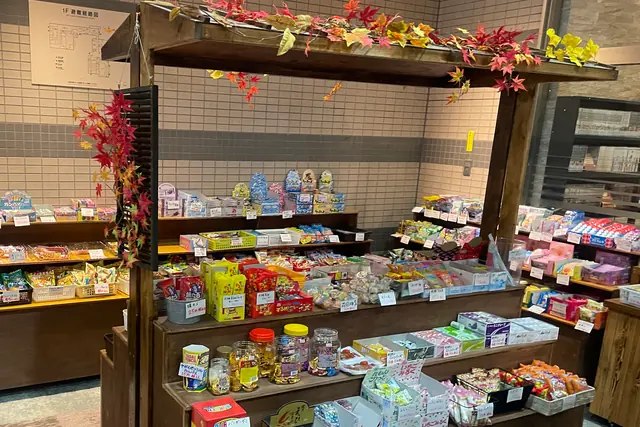 A wooden market-style stall with a small roof trimmed with artificial maple leaves, its tiered shelves packed with small packets of Japanese sweets and snacks