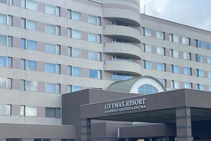 A dark entrance canopy on stone-clad pillars carrying the lettering LIVEMAX RESORT HAKODATE GREENPIA ONUMA, with the hotel block behind it