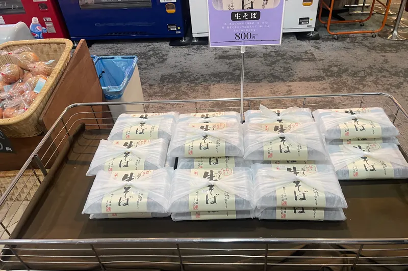 A wire trolley filled with flat packs of fresh buckwheat noodles under a handwritten sign giving a price of 800 yen including tax