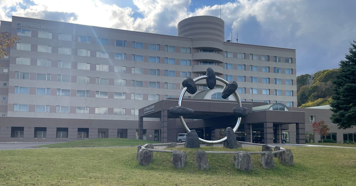 A long multi-storey hotel block with a round tower behind a lawn, with a large steel ring holding boulders standing in front of the entrance canopy