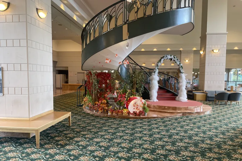 A hotel lobby with a green floral carpet, a display of red maple leaves, a stone lantern and a paper parasol beside a spiral staircase, and a white floral arch on a round red stage