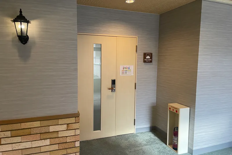 A dark wooden door in a pale corridor wall with a plaque beside it reading MAX CABIN ROOM 1-44 and a wall lamp above