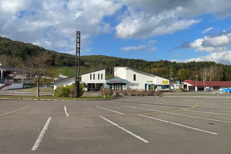 An empty car park in front of a white ticket building and a red-roofed lodge, with a grassy ski slope rising on the wooded hill behind them under a blue sky with clouds
